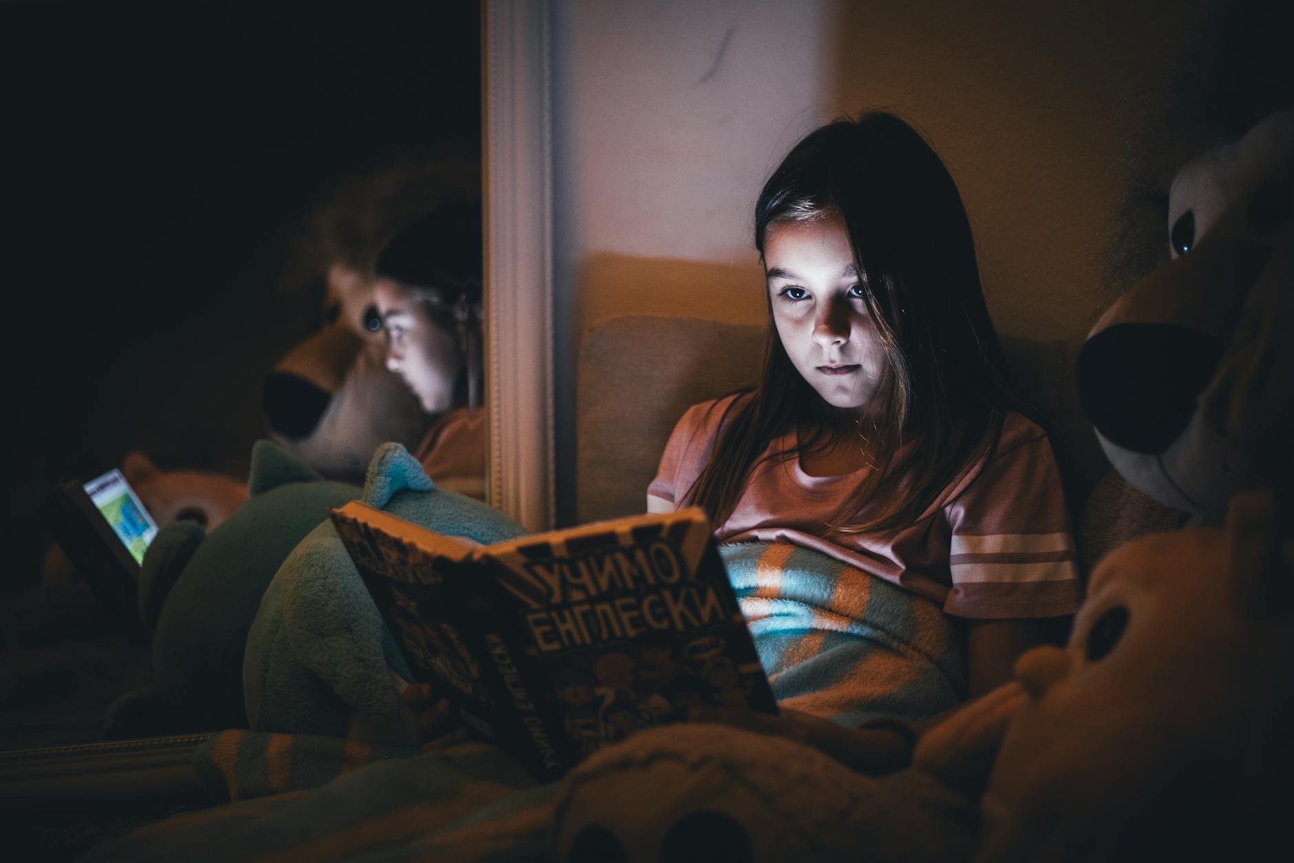 Dziewczynka czyta książkę obok świecącego tabletu wieczorem